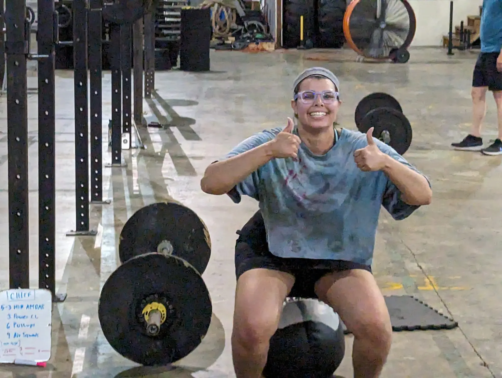 A smiling woman giving two thumbs-up while sitting on a medicine ball between barbell plates on the gym floor.