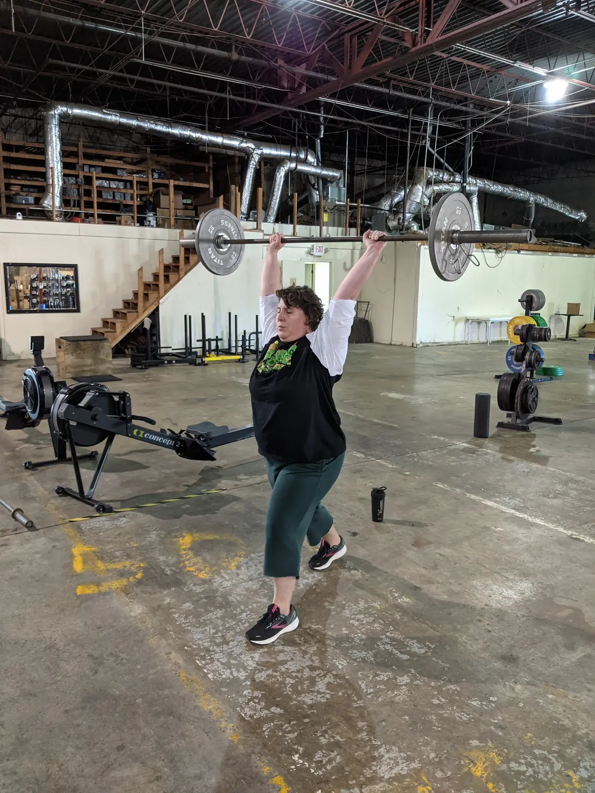 A woman holding a loaded barbell overhead while walking a lunge across the concrete floor of the Fit 901 warehouse gym.