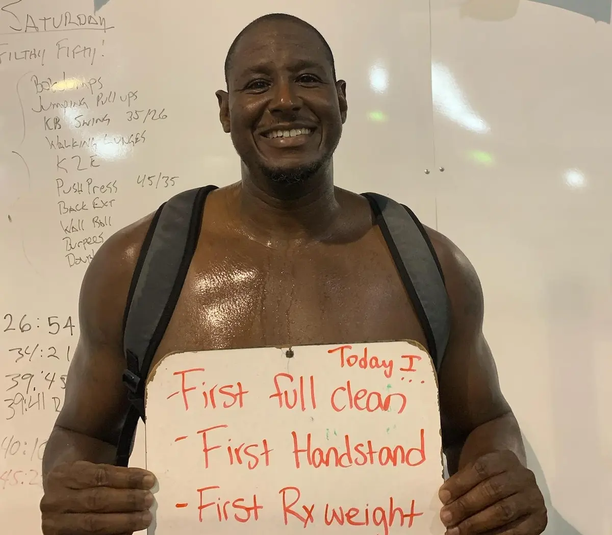 Chauncey, a Fit 901 client over 50, smiling and holding a board that reads: Today I: first full clean, first handstand, first Rx weight.