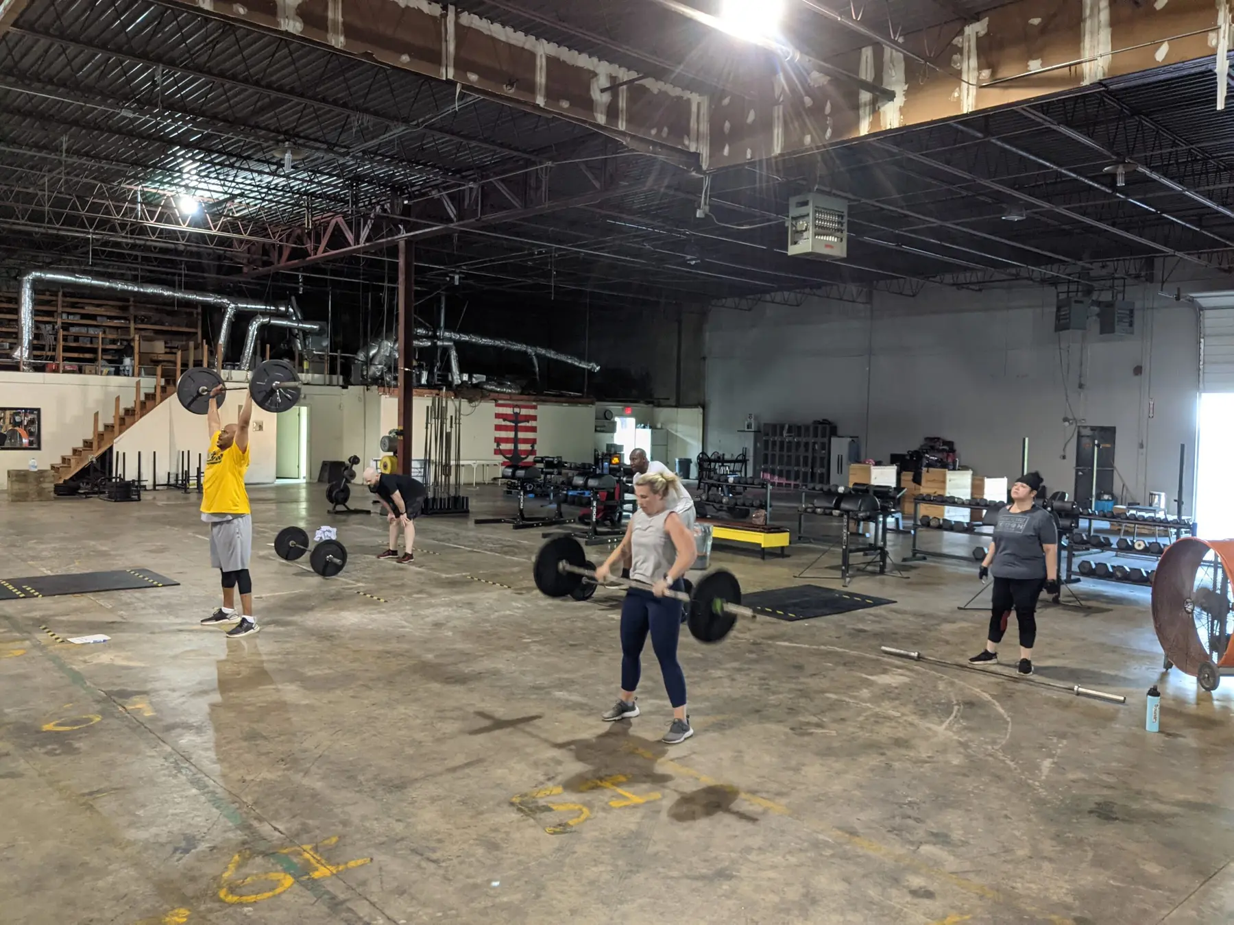 A Fit 901 session training with barbells across the open warehouse floor, one member pressing a barbell overhead, others mid-lift.