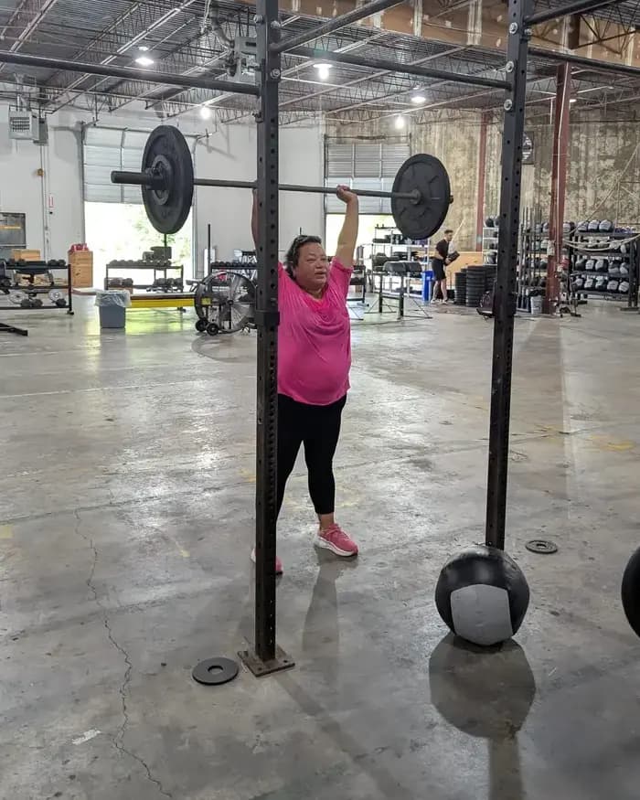 Diana, a Fit 901 client in her mid-50s, pressing a loaded barbell overhead on the concrete floor of the warehouse gym.