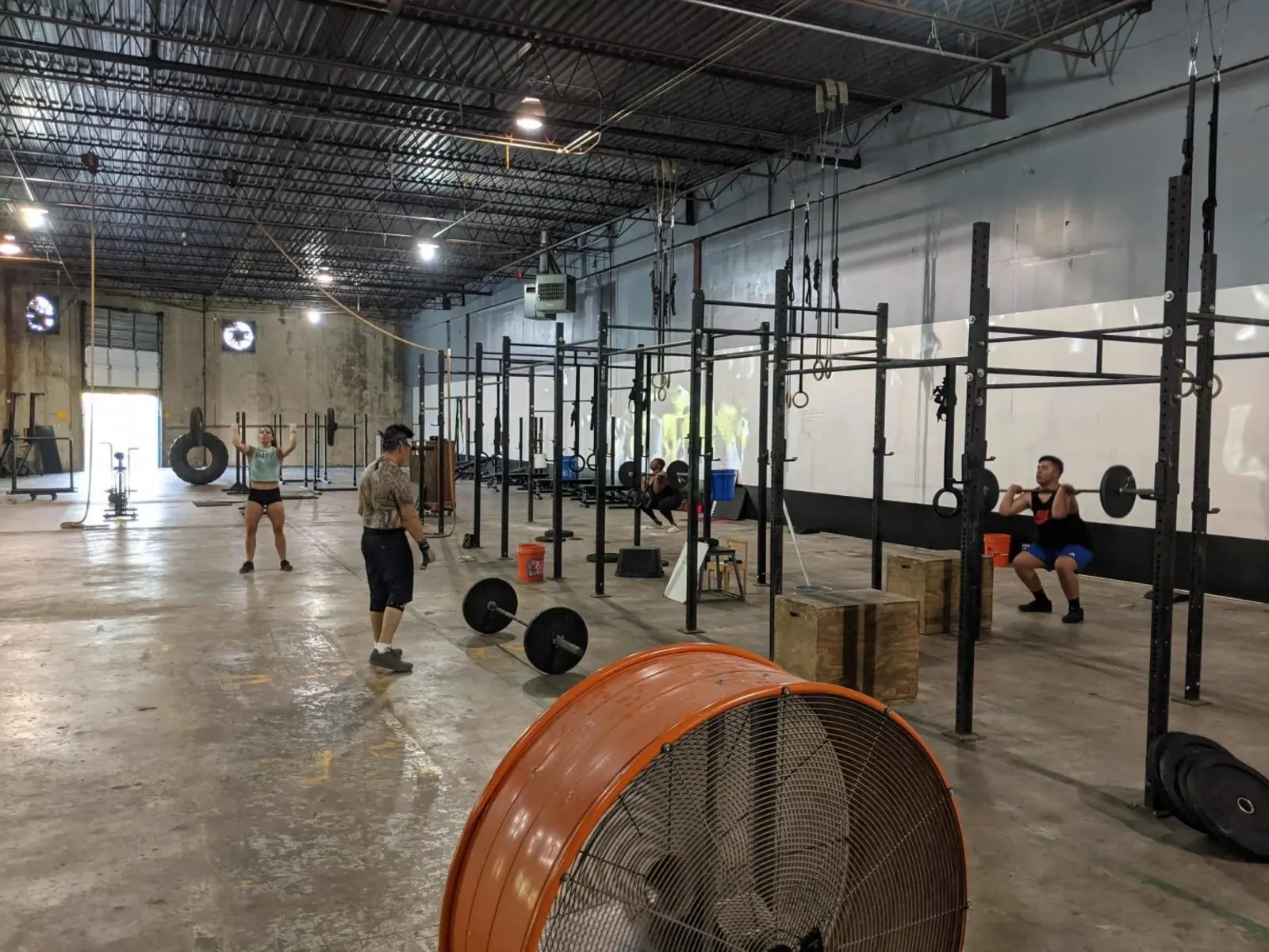 The Fit 901 training floor: bare concrete with lifting lines painted on it, an exposed steel deck ceiling, squat rigs along the wall, a large floor fan in the foreground, and members training. No mirrors.