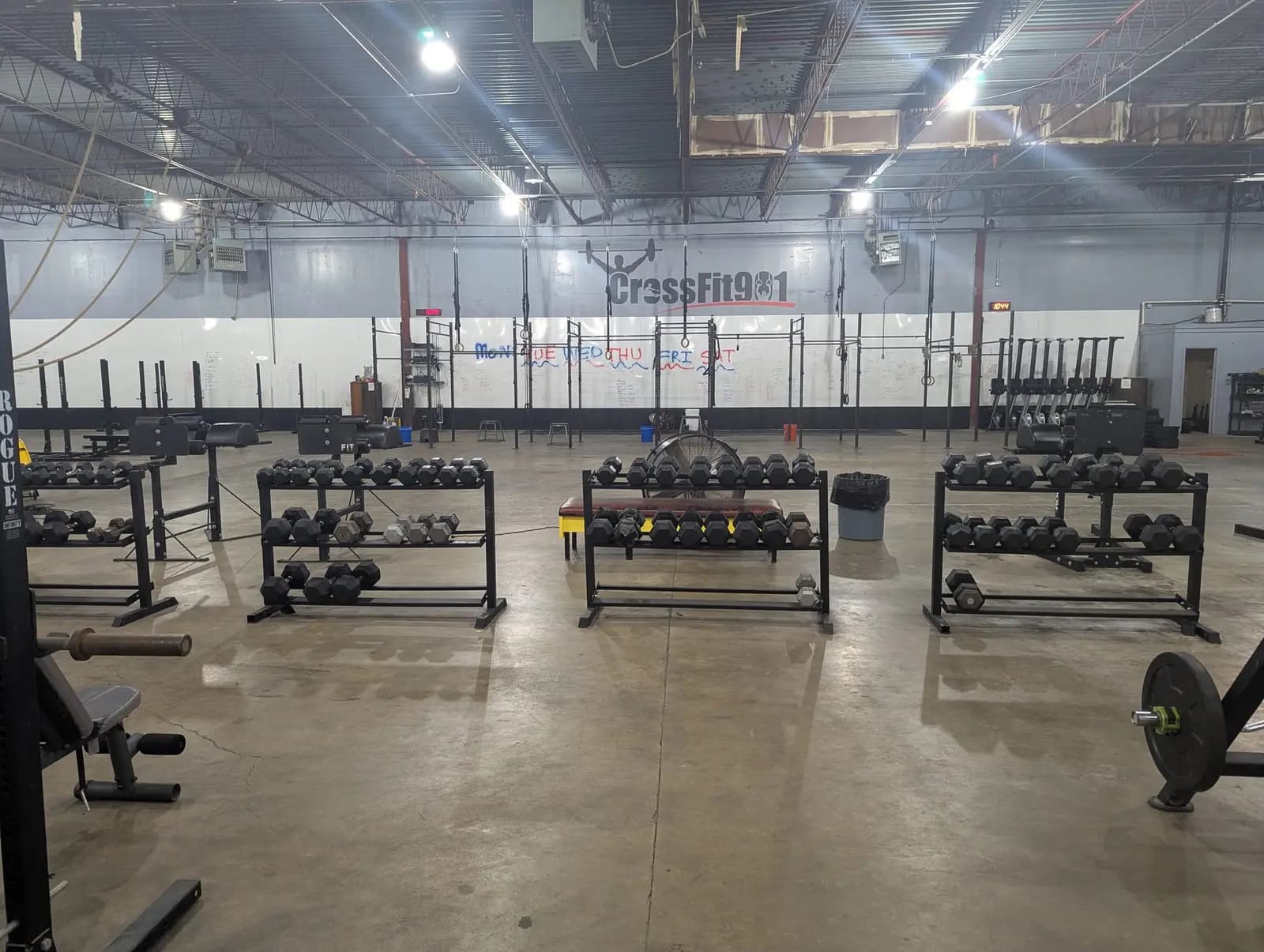 The Fit 901 training floor: an open warehouse with concrete floors, dumbbell racks, squat rigs, the week's programming written on a whiteboard along the wall, and the CrossFit 901 logo painted above it.