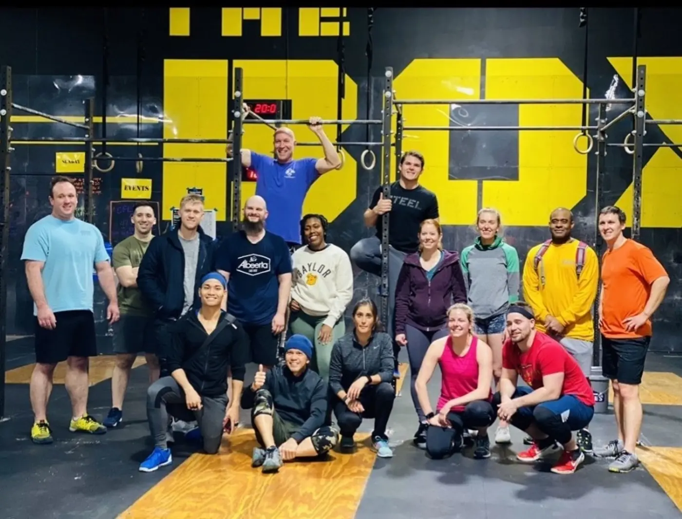 A large group of Fit 901 members posed together in the gym's previous building, in front of a black wall painted with a large yellow logo, some standing and some kneeling.