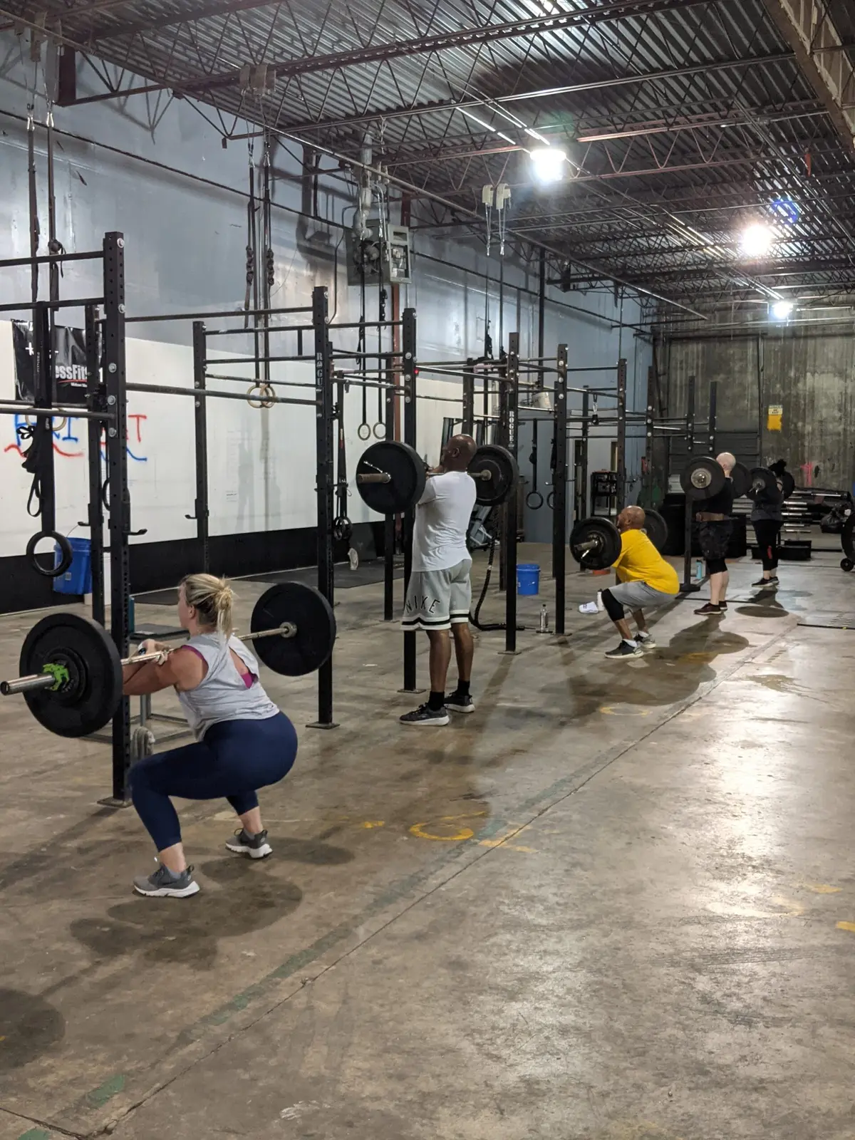 Four Fit 901 members back-squatting at separate racks along the wall, loaded barbells on their shoulders, on the concrete floor of the warehouse gym.