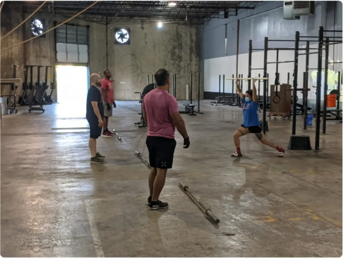 Fit 901 members warming up on the gym floor with empty barbells, one member overhead-squatting a barbell, coaches watching, roll-up loading door open at the back.