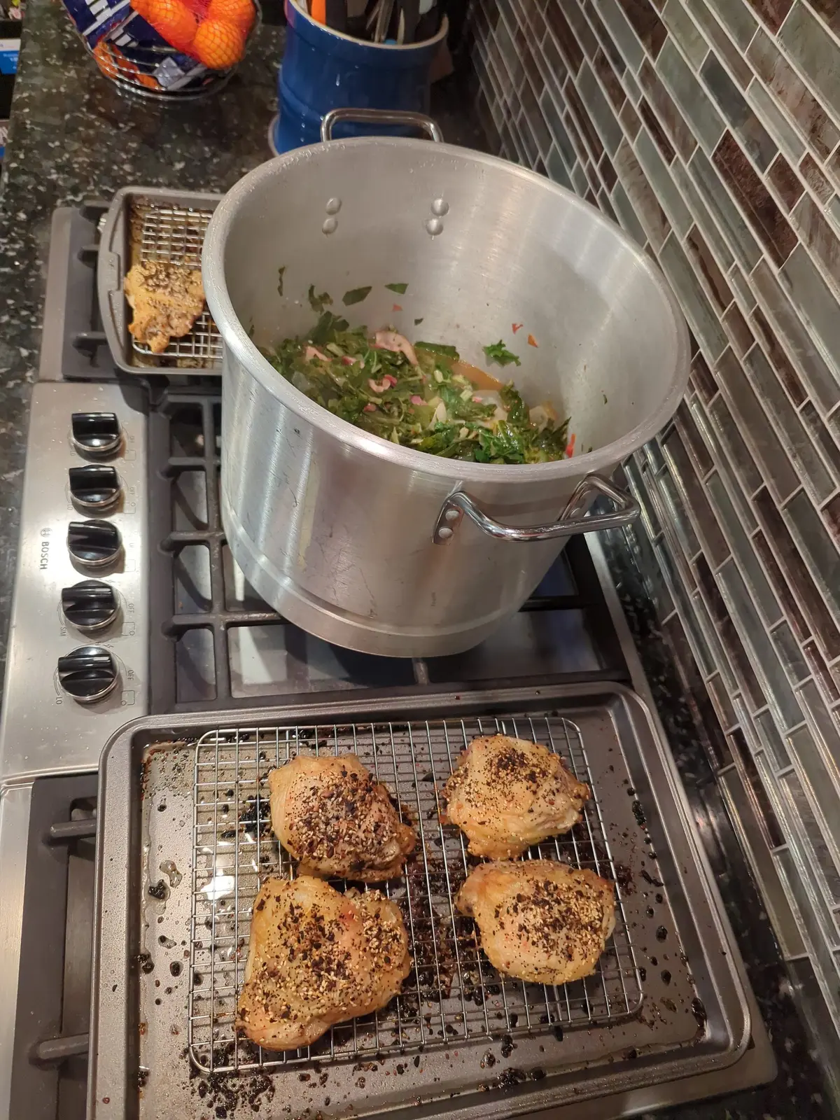 A home stove with a pot of greens simmering and a tray of seasoned chicken thighs resting on a rack.