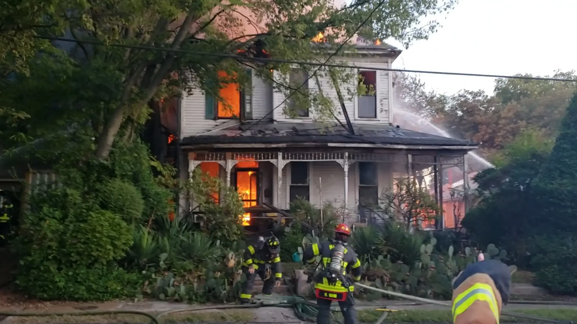Firefighters in turnout gear on the front lawn of a burning two-story house, fire showing from the upper windows and through the front porch. One of them, in a red helmet, is at the steps with a charged hoseline, about to go in.