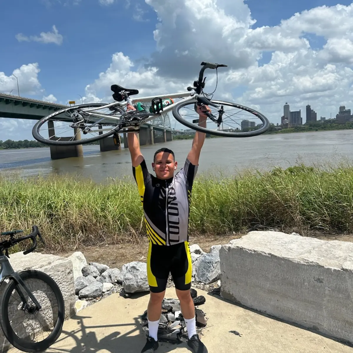 Alex holding his road bicycle above his head on the Memphis riverfront, the Hernando de Soto Bridge and downtown skyline behind him.