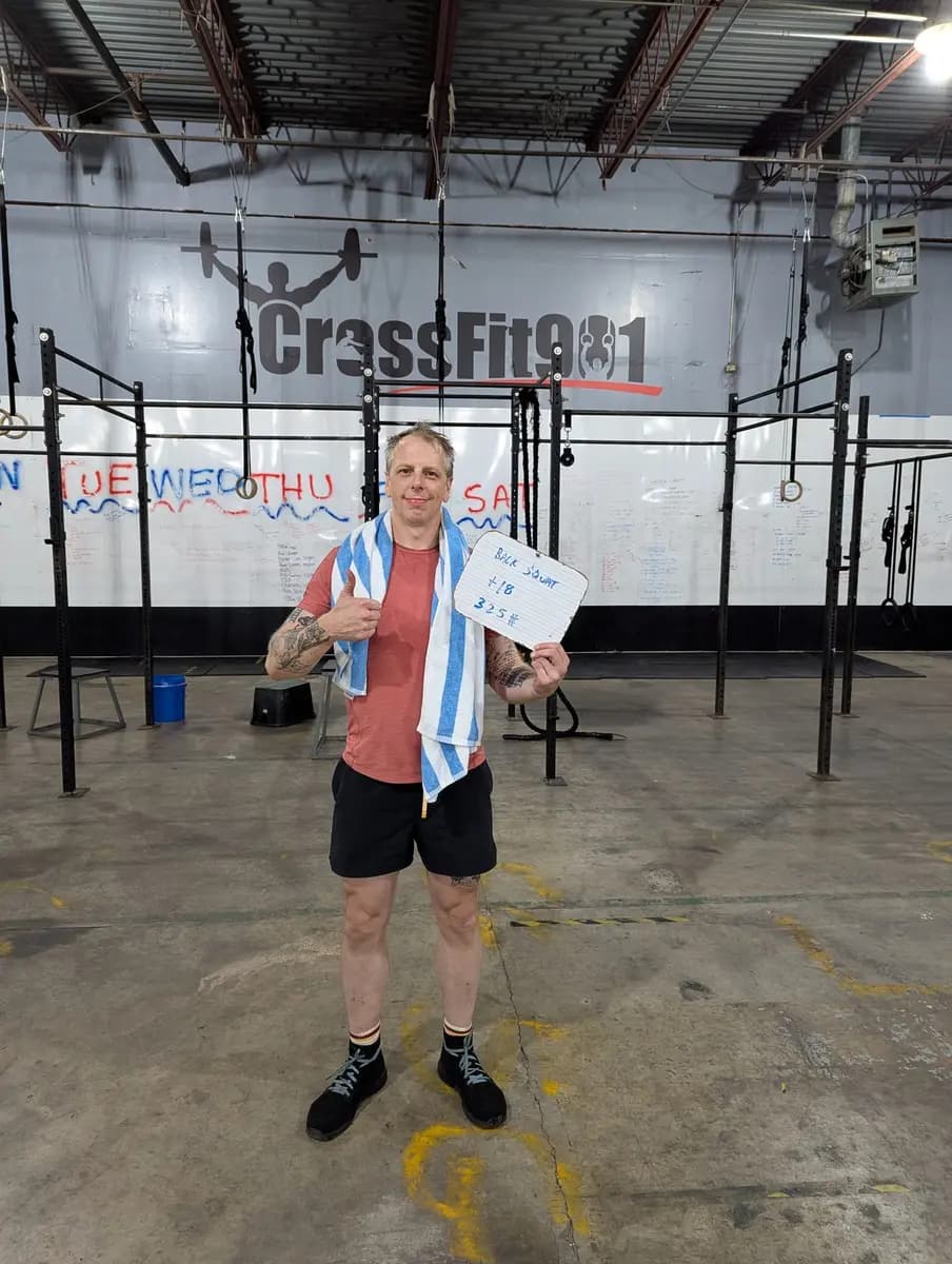 Matt holding a whiteboard reading BACK SQUAT +18, 325 pounds, in front of the CrossFit 901 wall logo.